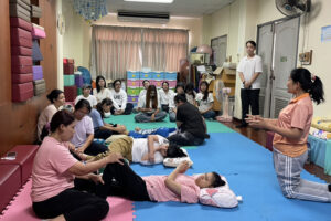 เจ้าหน้าที่นั่งอาธิบายกิจกรรม ให้กับผู้หญิงและผู้ชายนั่งใส่เสื้อสีขาวฟังในห้องกายภาพมีเด็กๆและผู้ปกครองฝึกน้องอยู่ด้วย