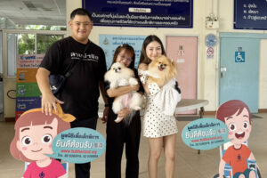 ผู้หญิง 2 คนยืนอุ่มหมาข้างผู้ชายด้านหน้ามีป้าย มูลนิธิเพื่อเด็กพิการ